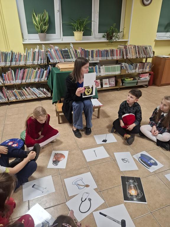 Bibliotekarka w niebieskiej bluzce i spodniach, czarnym swetrze siedzi na ławeczce, ma w ręku obrazek przedstawiający czapkę górnika. Grupa dzieci w kolorowych ubrania siedzi w kole. Na środku kola leżąc obrazki związane z pracą górnika. Za bibliotekarką jest stoli stolik z zielonym obrusem i regały z książkami.