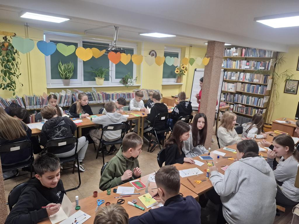 Grupa dzieci w kolorowych ubraniach siedzi przy dwóch rzędach ławek. Bibliotekarka w czerwonym swetrze stoi częściowo za słupem. Na stolikach leżą różne materiały potrzebne do zrobienia kartek świątecznych.