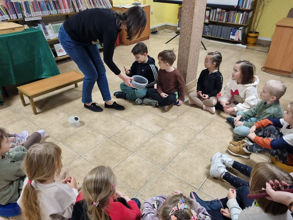 Bibliotekarka w czarnej bluzce i niebieskich spodniach trzyma pojemnik z węglem, pochyla się nad dziećmi i im go pokazuje. Drugi pojemniki swoi na podłodze. Grupa dzieci w kolorowych ubrania siedzi w kole, patrzą na węgiel.