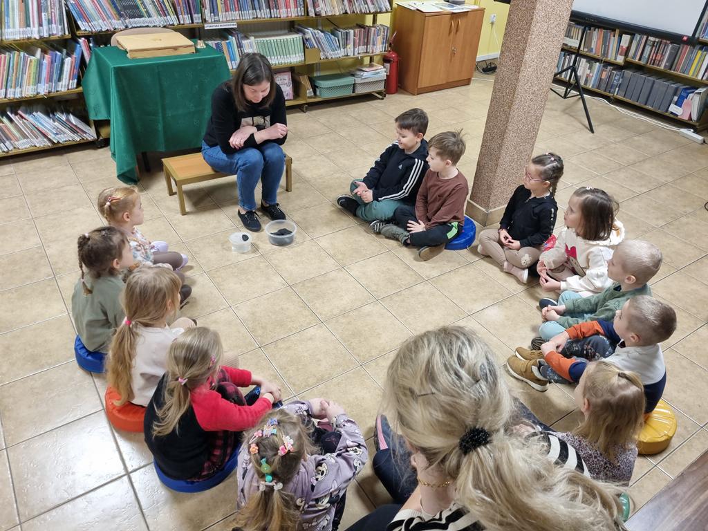 Bibliotekarka w czarnej bluzce i niebieskich spodniach siedzi na ławeczce, pochyla się nad dwoma pojemnikami z węgle. W jednym pojemniku jest woda. Grupa dzieci w kolorowych ubrania siedzi w kole, patrzą na węgiel. Za nimi na długiej ławce siedzi jedna panie nauczycielki.