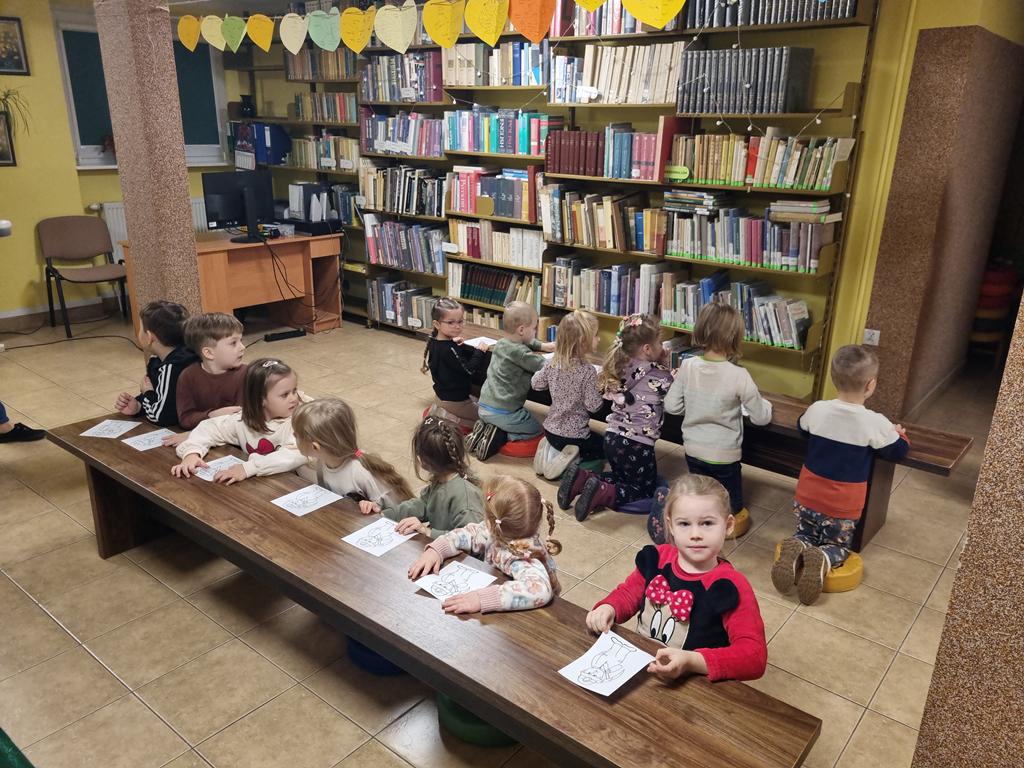 Grupa dzieci w kolorowych ubrania siedzi przy dwóch ławkach. Na ławkach rysunki górnika. W tle regały z książkami.