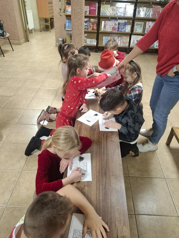 Dziewięcioro dzieci w kolorowych ubrania siedzi przy ławce i rozwiązuje labirynt. Kobiet w czerwonym swetrze i niebieskich spodniach stoi przy ławce i wręcza z pudełka ołówki. W tle są regały z książkami.