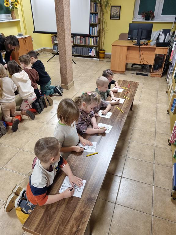 Grupa dzieci w kolorowych ubrania siedzi przy dwóch ławkach. Na ławkach rysunki górnika, kredki żółte i węgiel rysunkowy. Bibliotekarka w czarnej bluzce pochyla się nad dziećmi. Z tyłu biurko i biały ekran od rzutnika.