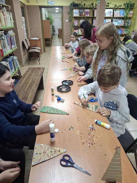 Grupa dzieci w kolorowych ubraniach siedzi przy ławkach i wykonują choinki z tekstury. Bibliotekarka w ciemno różowej bluzce schyla się nad dziećmi. Na stolikach kleje, nożyczki, tekturowe choinki, ozdoby.