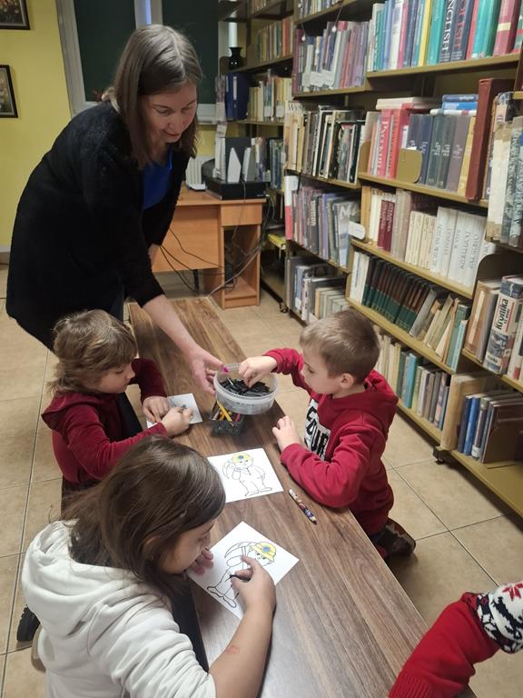 Troje dzieci w kolorowych ubrania siedzi przy ławce i maluje górnika węglem rysunkowym. Nad dziećmi nachyla się bibliotekarka, rozdaje węgiel rysunkowy. Na ławce są również kolorowe kredki. Po prawej stronie są regały z książkami.