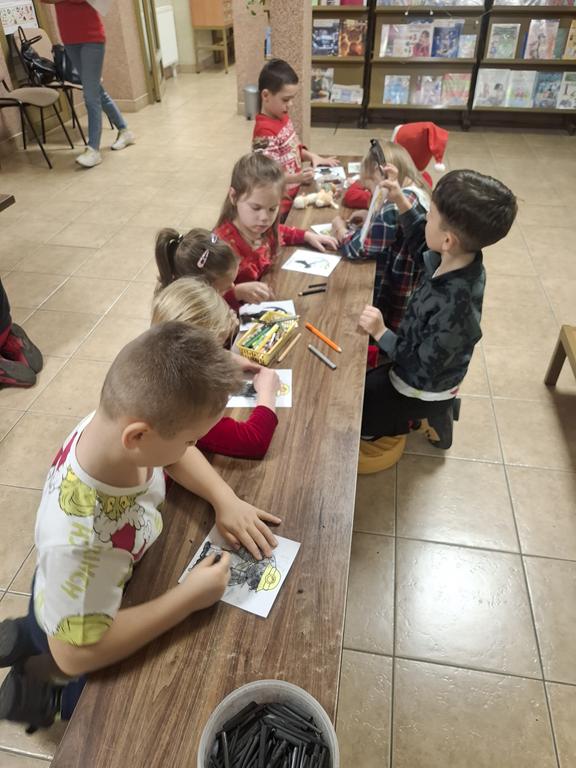 Grupa dzieci w kolorowych ubrania siedzi przy ławce i maluje górnika węglem rysunkowym. Na ławce są również kolorowe kredki. W tle są regały z książkami.