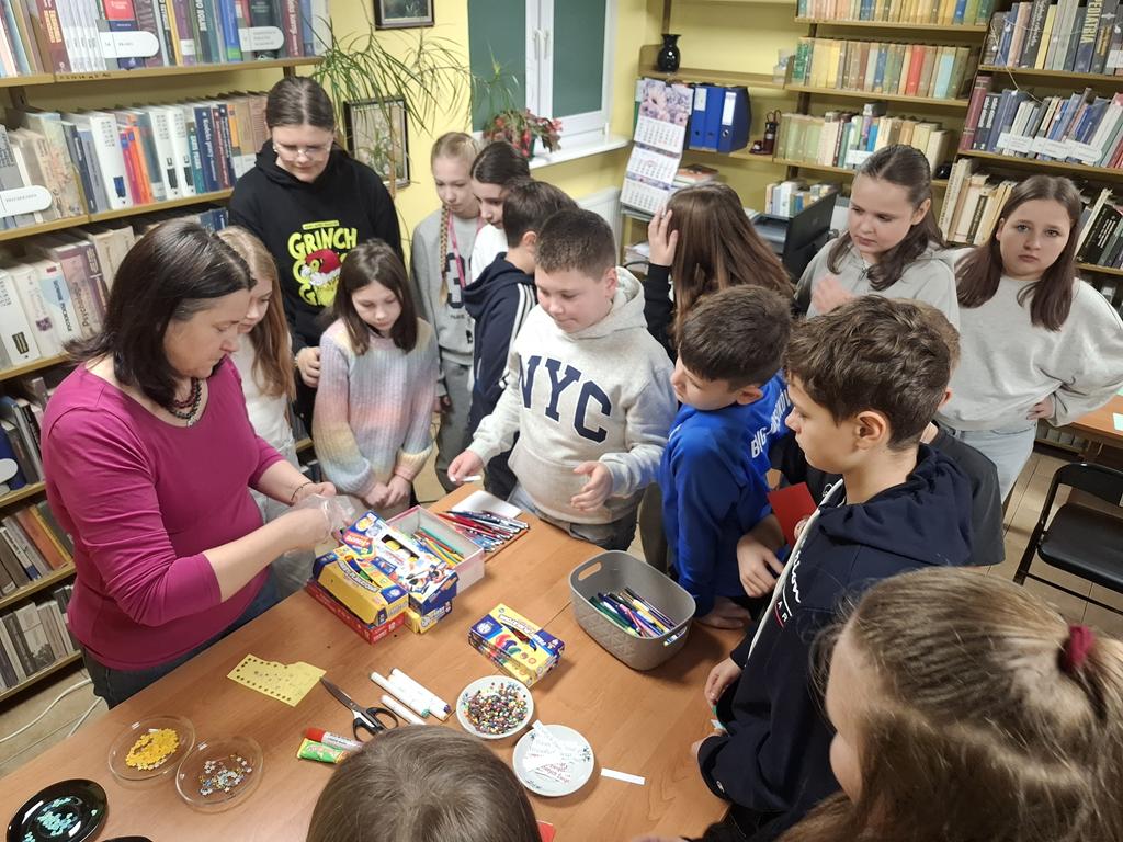 Przy stoliku z różnymi ozdobami stoi grupa dzieci w kolorowych ubraniach oraz bibliotekarka w ciemno różowej bluzce. Bibliotekarka wyjmuje coś z torebki, tłumaczy dzieciom co mają robić.
