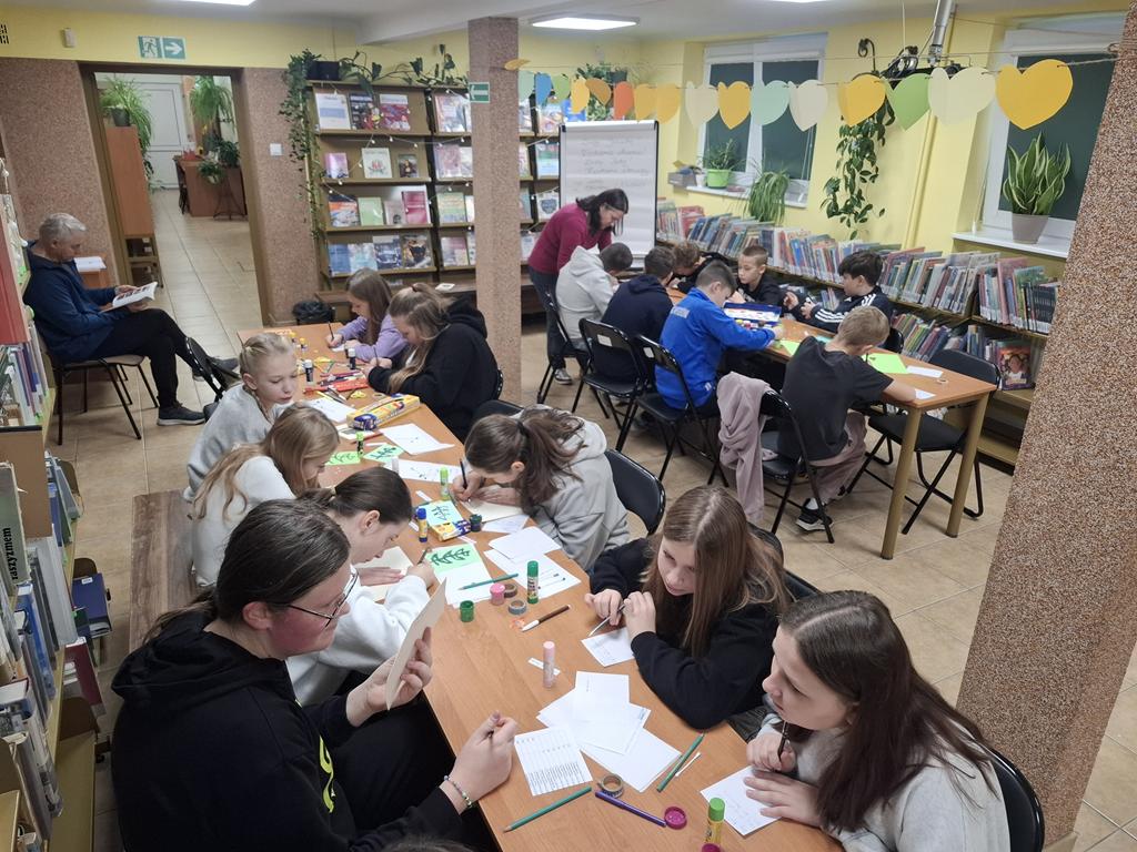 Grupa dzieci w kolorowych ubraniach siedzi przy dwóch rzędach stolików. Na stolikach leżą różne rzeczy do wykonania kartek świątecznych. Bibliotekarka w ciemno różowej bluzce schyla się nad dziećmi. Za dziećmi siedzi pan nauczyciel w ciemnych ubraniach.