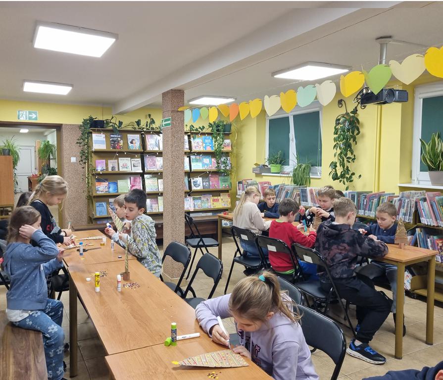 Grupa dzieci w kolorowych ubraniach siedzi przy dwóch rzędach ławek. Wykonują choinki z tektury i innych potrzebnych materiałów leżących na ławkach.