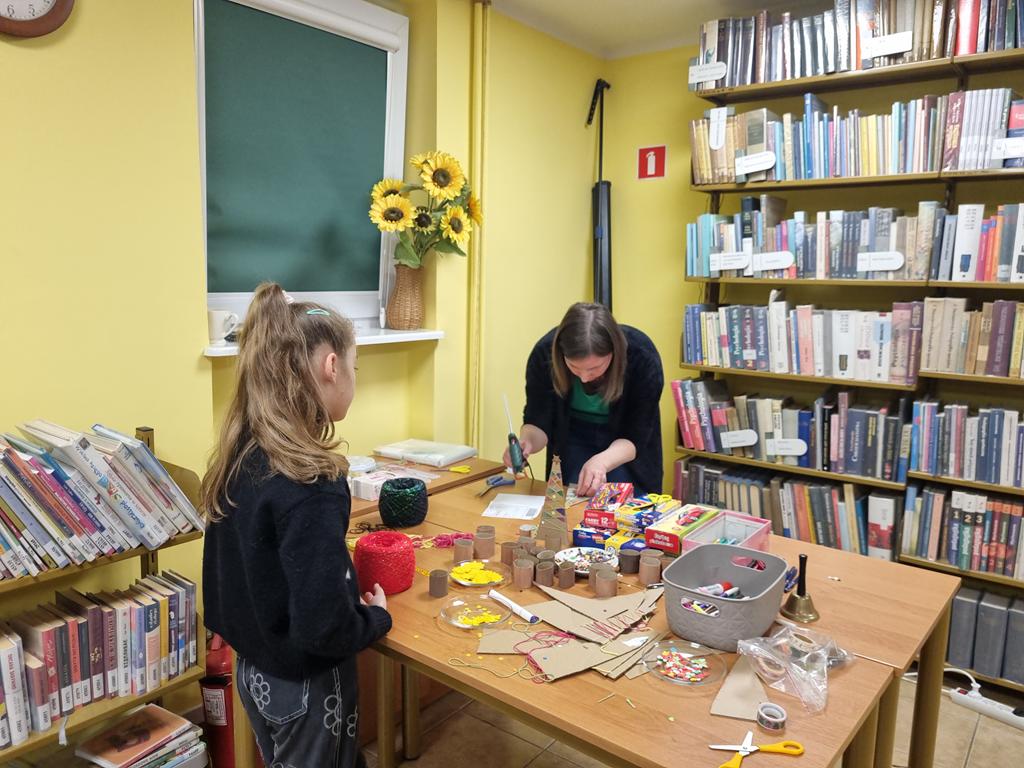 Bibliotekarka w zielonej bluzce, czarnym swetrze schyla się nad stolikiem. Używa kleju na gorąco. Obok stolika stoi dziewczynka w czarnym swetrze, niebieskich spodniach. Na stoliku są materiały potrzebne do zrobienia choinki. W tle regały z książkami.