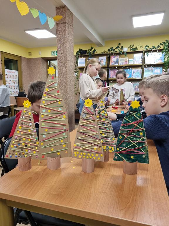 Pięć choinek wykonanych z kolorowego papieru i tektury. W tle dzieci w kolorowych ubraniach.