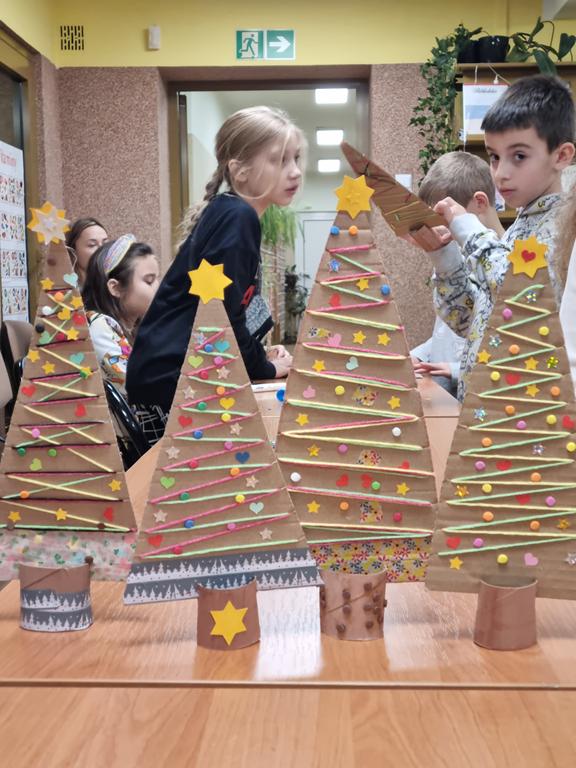 Cztery choinki wykonane z kolorowego papieru i tektury. W tle dzieci w kolorowych ubraniach.