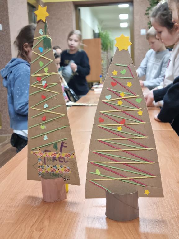 Dwie choinki wykonane z kolorowego papieru i tektury. W tle dzieci w kolorowych ubraniach.