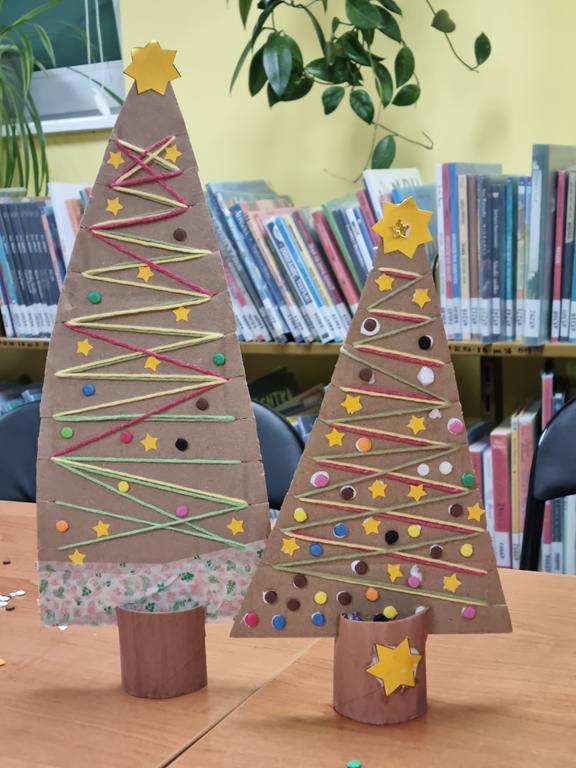 Dwie choinki wykonane z kolorowego papieru i tektury. W tle regały z książkami.