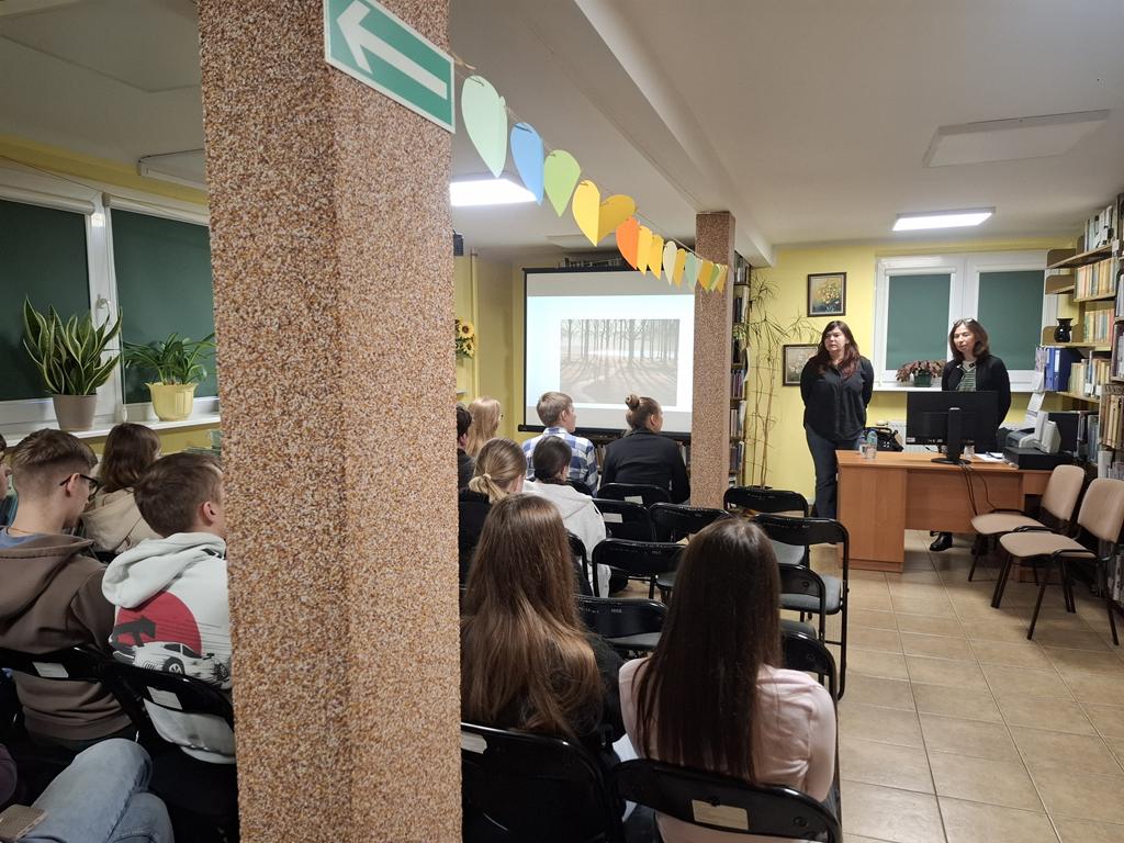 Grupa młodzieży siedzi na krzesłach. Przed nimi stoją za biurkiem dwie kobiety w ciemnych ubraniach. Obok kobiet stoi ekran rzutnika, na którym jest wyświetlana prezentacja dotycząca depresji.