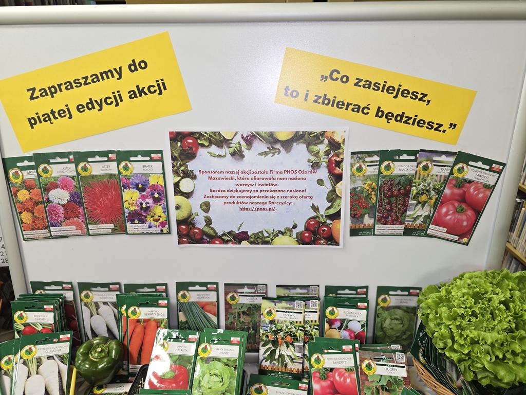 Na tablicy jest nazwa akcji, nazwa sponsora oraz nasiona warzyw i kwiatów. Przed tablicą leżą na stoliku torebki z nasionami.