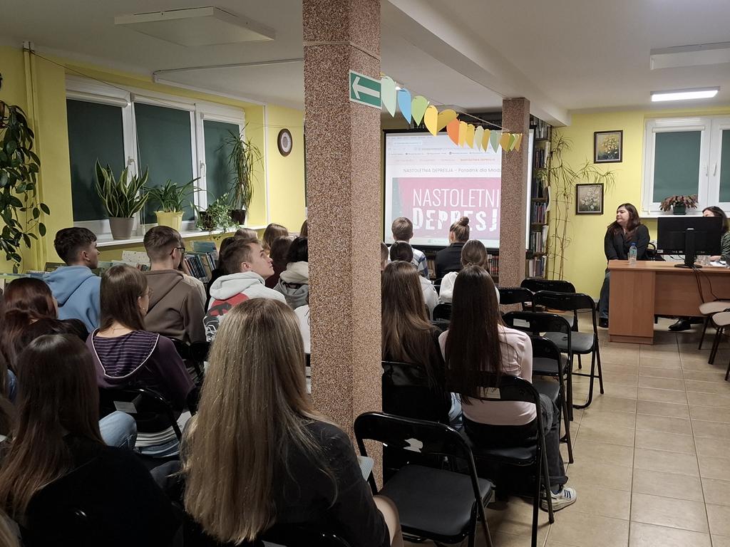 Grupa młodzieży siedzi na krzesłach. Przed nimi siedzą za biurkiem dwie kobiety w ciemnych ubraniach. Obok kobiet stoi ekran rzutnika, na którym jest wyświetlana prezentacja dotycząca depresji.