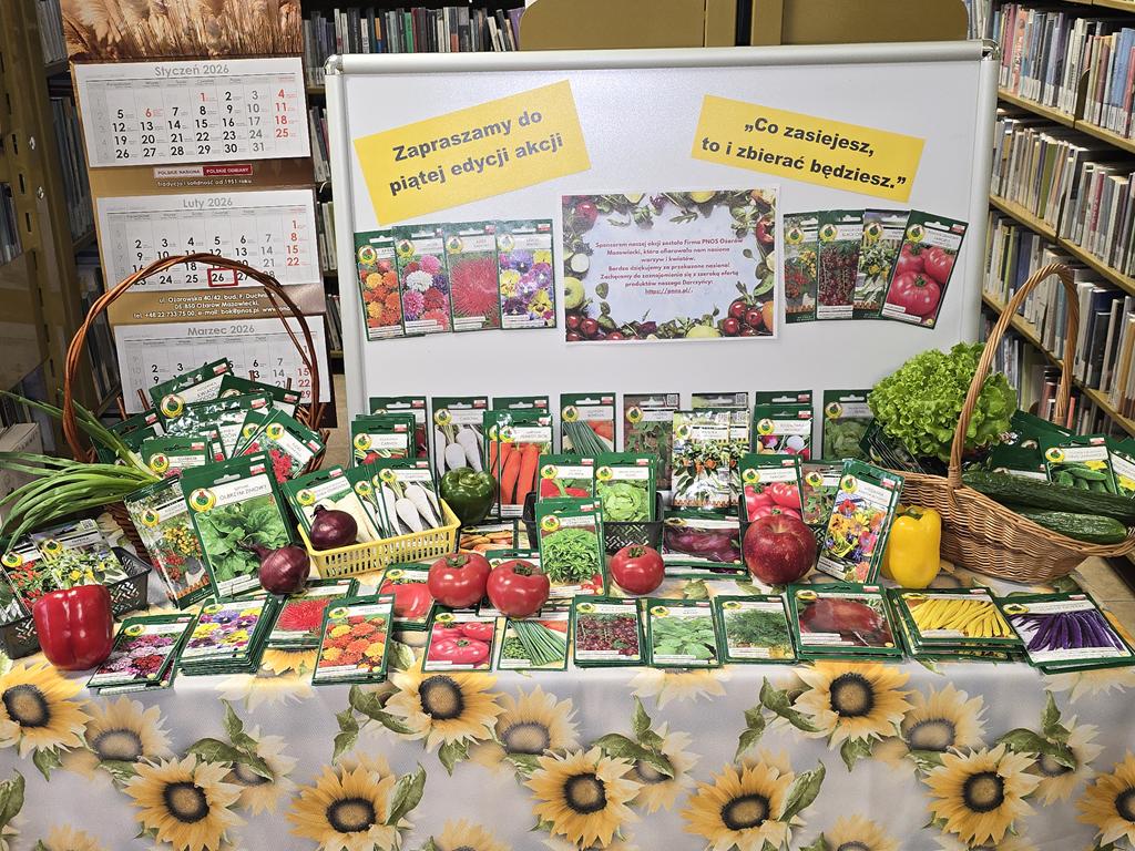 Na stoliku z obrusem w żółte kwiaty leżą torebki z nasionami warzyw i kwiatów. Pomiędzy nimi są trzy pomidory, jabłko, dwie papryki. Po prawej stronie stoi koszyk z nasionami ogórków, dwoma ogórkami i sałata. Po lewej stroni stoi koszyk z nasionami kwiatów i cebulą dymką. Z tyłu jest tablica z nazwą akcji, nazwą sponsora. Po lewej stronie tablicy wisi kalendarz sponsora.