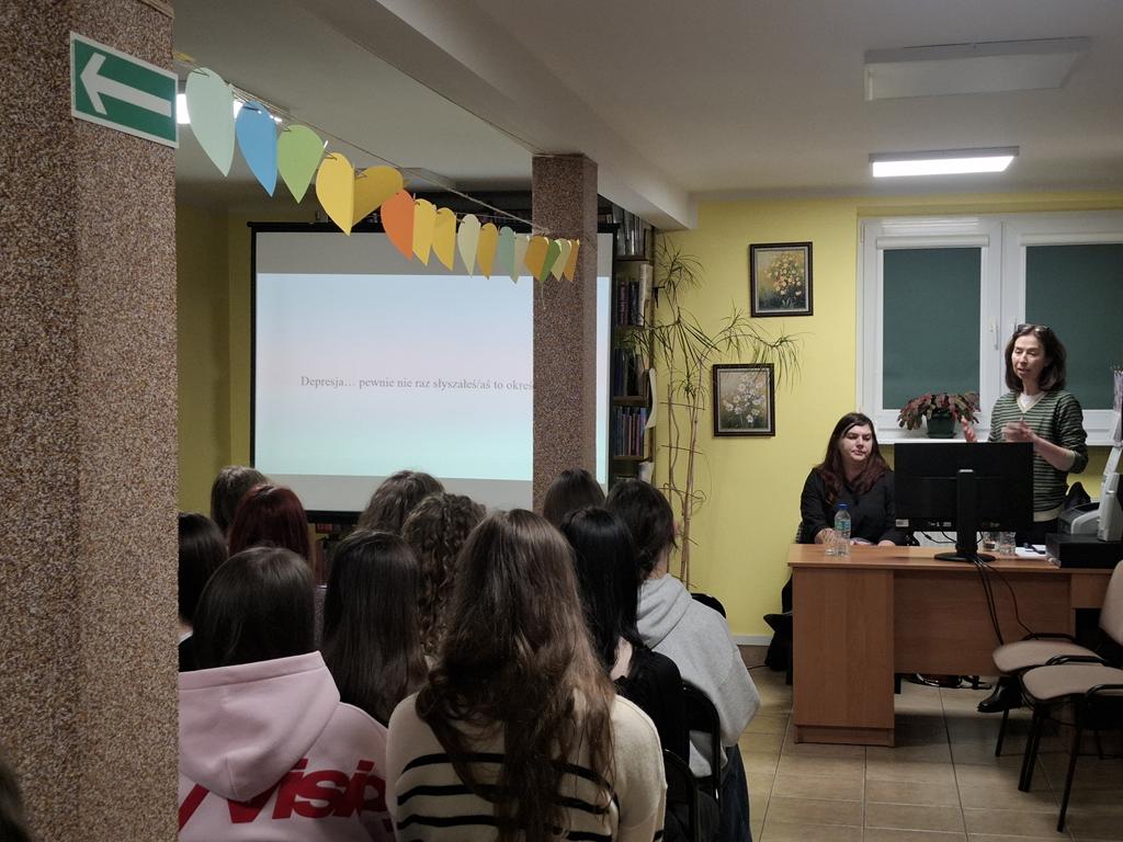 Grupa młodzieży siedzi na krzesłach. Przed nimi za biurkiem stoi jedna kobieta, druga siedzi. Obok kobiet stoi ekran rzutnika, na którym jest wyświetlana prezentacja dotycząca depresji.