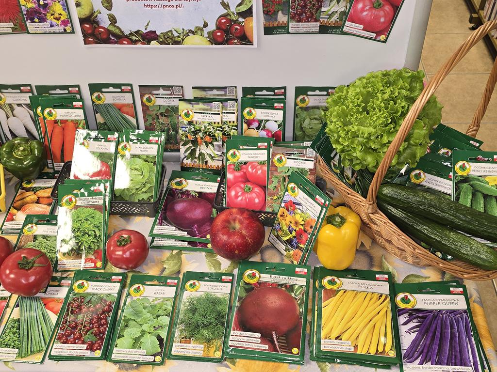 Na stoliku leżą torebki z nasionami warzyw i kwiatów. Pomiędzy nimi są dwa pomidory, jabłko, dwie papryki. Po prawej stronie stoi koszyk z nasionami ogórków, dwoma ogórkami i sałata.