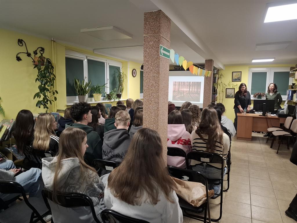 Grupa młodzieży siedzi na krzesłach. Przed nimi stoją za biurkiem dwie kobiety w ciemnych ubraniach. Obok kobiet stoi ekran rzutnika, na którym jest wyświetlana prezentacja dotycząca depresji.