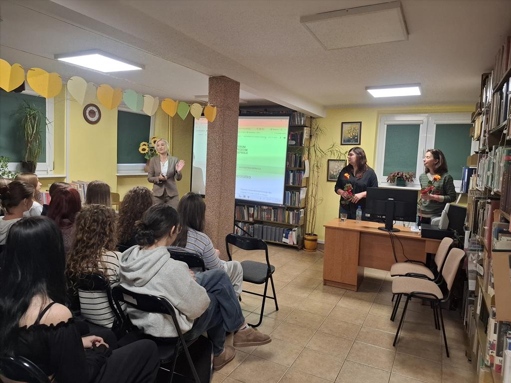 Grupa młodzieży siedzi na krzesłach. Przed nimi stoją za biurkiem dwie kobiety w ciemnych ubraniach, trzymają po jednej róży. Obok kobiet stoi ekran rzutnika, na którym jest wyświetlana prezentacja dotycząca depresji. Po lewej stroni rzutnika stoi trzecia kobieta w jasnych ubraniach.