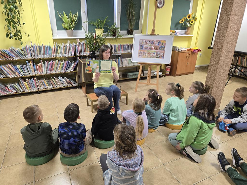 Bibliotekarka w żółtej bluzce i niebieskich spodniach siedzi na środku sali i trzyma książkę. Za nią jest stolik z zielonym obrusem, na którym stoi wazon z palmą i bukszpanem oraz 4 pisanki. Przed bibliotekarką siedzi grupa dzieci w kolorowych ubraniach. Po prawej stronie bibliotekarki jest tablica z plakatem dotyczącym Świąt Wielkanocnych. W tle są regały z książkami.