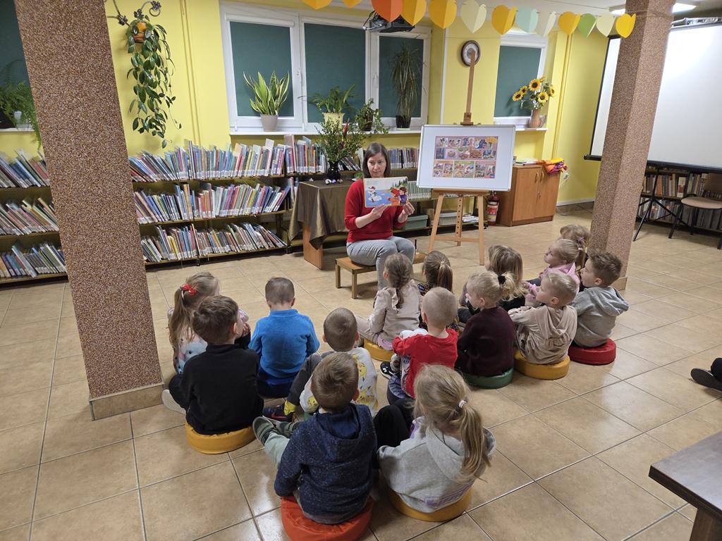 Bibliotekarka w czerwonej bluzce i szarych spodniach siedzi na ławeczce i trzyma książkę. Za nią stoi stoli z zielonym obrusem, na którym jest wazon z palmą, bukszpanem oraz 4 pisanki. Obok stolika jest tablica z plakatem dotyczącym świąt Wielkanocnych. Przed kobietą siedzi grupa dzieci w kolorowych ubraniach. W tle są regały z książkami.