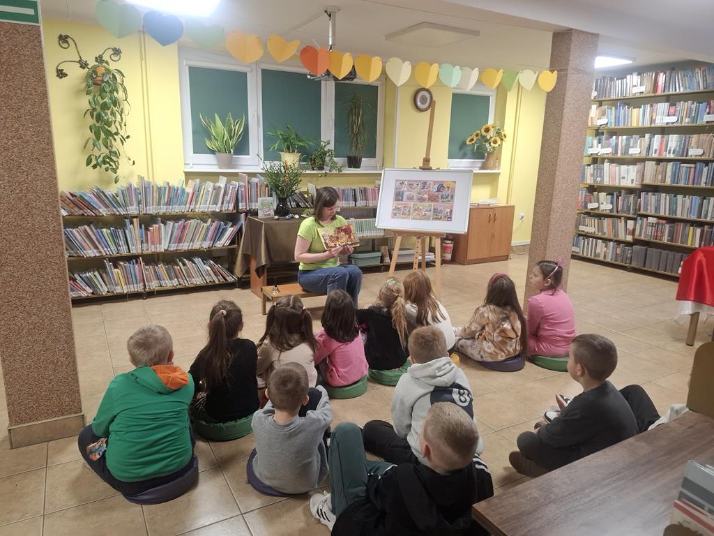 Bibliotekarka w zielonej bluzce i niebieskich spodniach siedzi na środku sali i trzyma książkę. Za nią jest stolik z zielonym obrusem, na którym stoi wazon z palmą i bukszpanem, 4 pisanki, książka dotycząca Świąt Wielkanocnych. Przed bibliotekarką siedzi grupa dzieci w kolorowych ubraniach. Po prawej stronie bibliotekarki jest tablica z plakatem dotyczącym Świąt Wielkanocnych. W tle są regały z książkami.