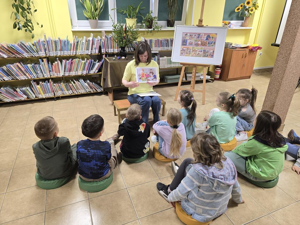 Bibliotekarka w żółtej bluzce i niebieskich spodniach siedzi na środku sali i trzyma obrazek dotyczący Świąt Wielkanocnych. Za nią jest stolik z zielonym obrusem, na którym stoi wazon z palmą i bukszpanem oraz 4 pisanki. Przed bibliotekarką siedzi grupa dzieci w kolorowych ubraniach. Po prawej stronie bibliotekarki jest tablica z plakatem dotyczącym świąt. W tle są regały z książkami.