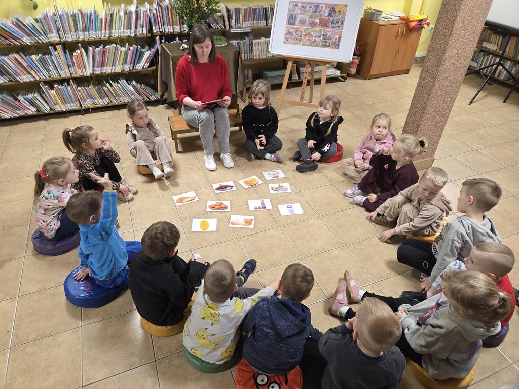 Bibliotekarka w czerwonej bluzce i szarych spodniach siedzi na ławeczce i trzyma podkładkę z zagadkami. W kręgu razem z kobietą siedzi grupa dzieci w kolorowych ubraniach. Za nią stoi stoli z zielonym obrusem, na którym jest wazon z palmą, bukszpanem oraz 4 pisanki. Obok stolika jest tablica z plakatem dotyczącym świąt Wielkanocnych. W tle są regały z książkami.