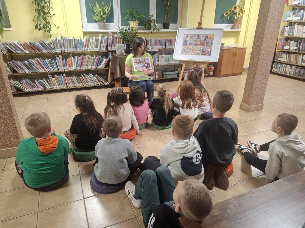 Bibliotekarka w zielonej bluzce i niebieskich spodniach siedzi na środku sali i trzyma obrazek dotyczący Świąt Wielkanocnych. Za nią jest stolik z zielonym obrusem, na którym stoi wazon z palmą i bukszpanem, 4 pisanki, książka dotycząca Świąt Wielkanocnych. Przed bibliotekarką siedzi grupa dzieci w kolorowych ubraniach. Po prawej stronie bibliotekarki jest tablica z plakatem dotyczącym świąt. W tle są regały z książkami.