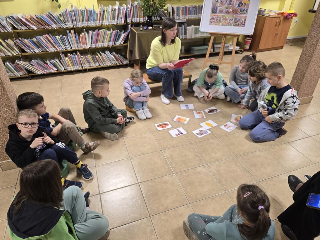 Bibliotekarka w żółtej bluzce i niebieskich spodniach siedzi w kręgu i trzyma podkładkę z zagadkami. Razem z nią w kręgu siedzi grupa dzieci w kolorowych ubraniach. W środku kręgu leżą ilustracje związane ze świętami. Za bibliotekarką jest stolik z zielonym obrusem, na którym stoi wazon z palmą i bukszpanem oraz 4 pisanki. Po prawej stronie bibliotekarki jest tablica z plakatem dotyczącym Świąt Wielkanocnych. W tle są regały z książkami.