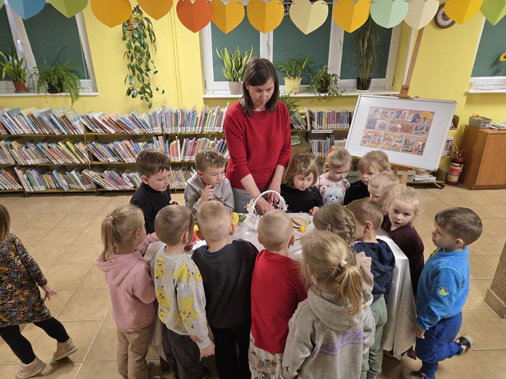 Bibliotekarka w czerwonej bluzce i szarych spodniach stoi przy stoliku z białym obrusem. Na stoliku leżą: koszyk, jajko, baranek, jabłko, cytryna, chrzań w słoiku, kiełbaska, chleb, solniczka i pieprzniczka, ozdoby do koszyka, papryka, przyprawy. Dokoła stolika stoją dzieci w kolorowych ubraniach.