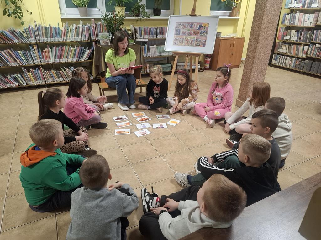 Bibliotekarka w zielonej bluzce i niebieskich spodniach siedzi w kręgu i trzyma podkładkę z zagadkami. Razem z nią w kręgu siedzi grupa dzieci w kolorowych ubraniach. W środku kręgu leżą ilustracje związane ze świętami. Za bibliotekarką jest stolik z zielonym obrusem, na którym stoi wazon z palmą i bukszpanem, 4 pisanki, książka dotycząca Świąt Wielkanocnych. Po prawej stronie bibliotekarki jest tablica z plakatem dotyczącym Świąt Wielkanocnych. W tle są regały z książkami.