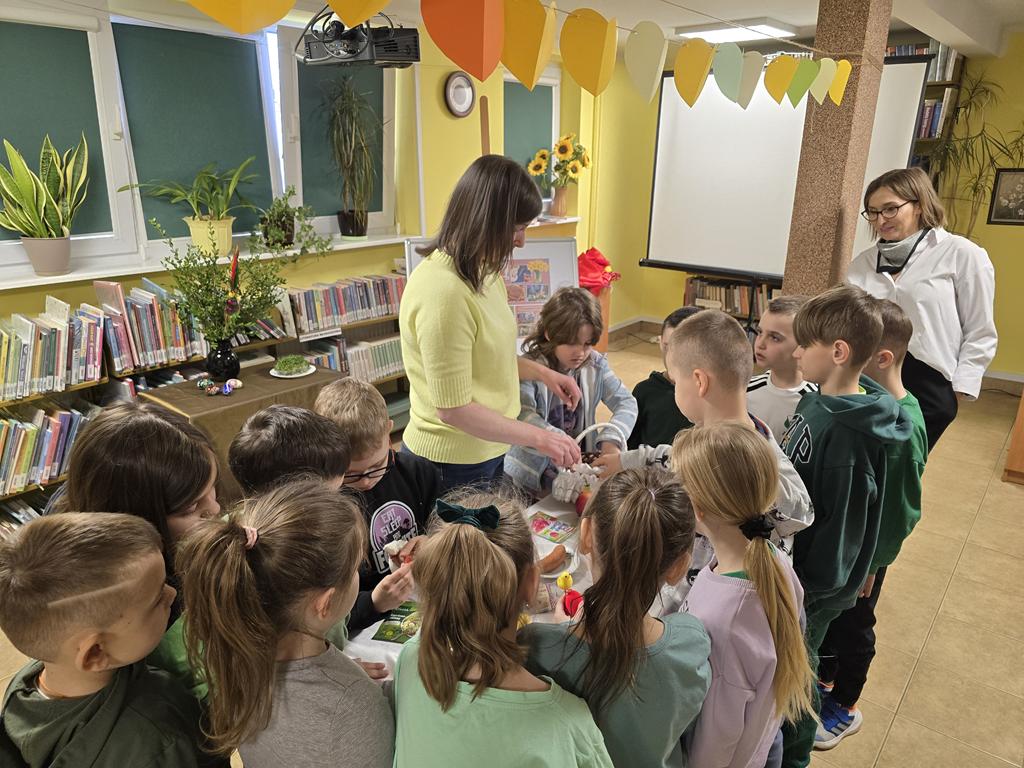 Pani bibliotekarka w żółtej bluzce i niebieski spodniach stoi przy stoliku z białym obrusem. Na stoliku leżą: koszyk, jajko, baranek, jabłko, cytryna, chrzań w słoiku, kiełbaska, chleb, solniczka i pieprzniczka, ozdoby do koszyka, papryka, przyprawy. Dokoła stolika stoją dzieci w kolorowych ubraniach.