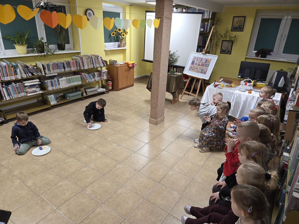 Dwóch chłopiec kuca przed talerzami z pisankami. Kręcą pisankami. Przed nimi siedzi grupa dzieci w kolorowych ubraniach. W tle jest ekran od rzutnika i regały z książkami.