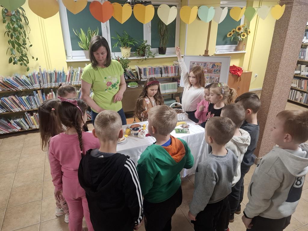 Pani bibliotekarka w zielonej bluzce i niebieski spodniach stoi przy stoliku z białym obrusem. Na stoliku leżą: koszyk, jajko, baranek, jabłko, cytryna, chrzań w słoiku, kiełbaska, chleb, solniczka i pieprzniczka, ozdoby do koszyka, papryka, przyprawy. Dokoła stolika stoją dzieci w kolorowych ubraniach.