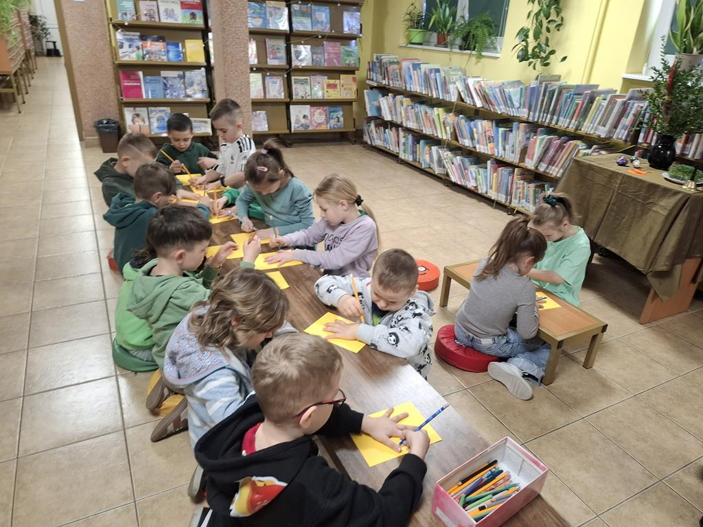 Dziesięcioro dzieci w kolorowych ubraniach siedzi przy ławce. Dwie dziewczynki siedzą przy inne ławeczce. Na ławce leżą żółte kartki, ołówki. Dzieci odrysowują rękę na kartce. W tle są regały z książkami.