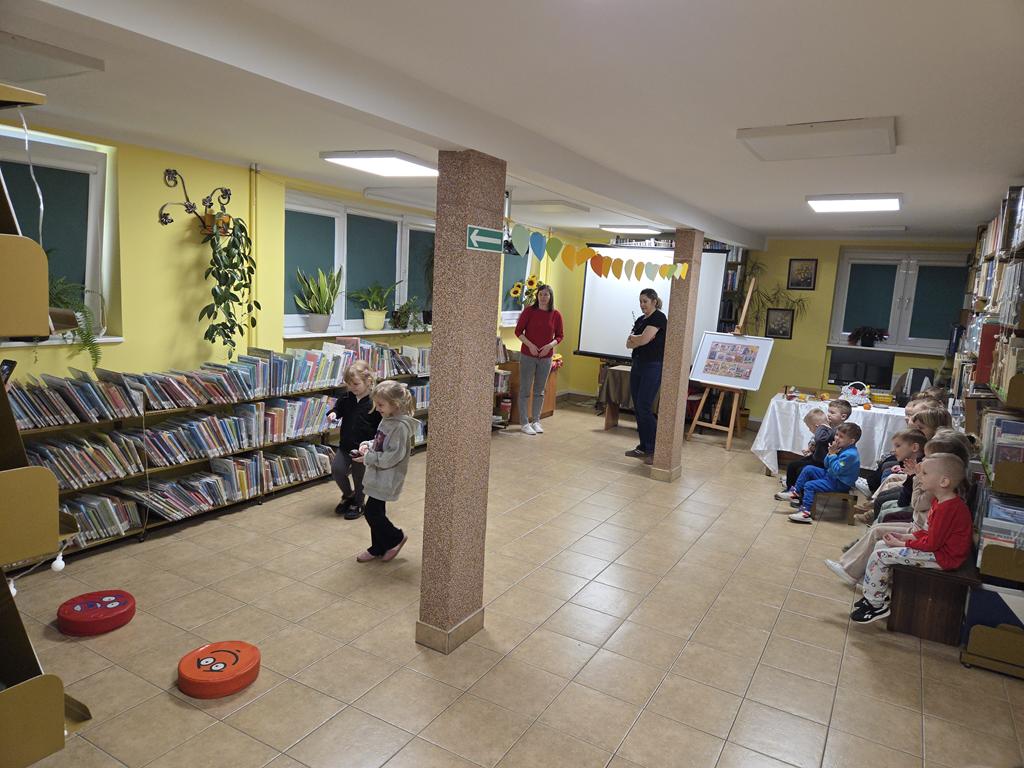 Dwie dziewczyna w kolorowych ubraniach trzymają w rękach łyżkę z pisanką, biorą udział z zawodach. Po ich prawej stronie siedzi grupa dzieci w kolorowych ubraniach. Przy słupie stoi pani nauczycielka w czarnych ubraniach, na przeciwko niej stoi bibliotekarka w czerwonej bluzce i szarych spodniach. W tle jest ekran od rzutnika i regały z książkami.