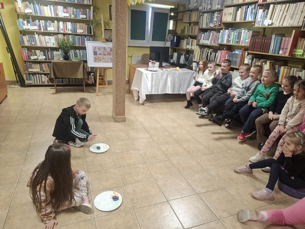 Chłopiec i dziewczynka w kolorowych ubraniach kucają przed talerzami i kręcą pisanką. Po ich prawej stronie siedzi grupa dzieci w kolorowych ubraniach. W tle jest ekran od rzutnika.