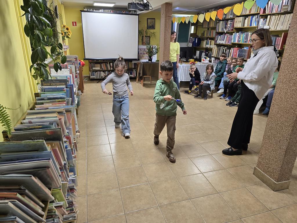 Chłopiec i dziewczyna w kolorowych ubraniach trzymają w rękach łyżkę z pisanką, biorą udział z zawodach. Po ich prawej stronie siedzi grupa dzieci w kolorowych ubraniach. Przy słupie stoi Pani nauczycielka w białej bluzce i czarnych spodniach. W tle jest ekran od rzutnika.