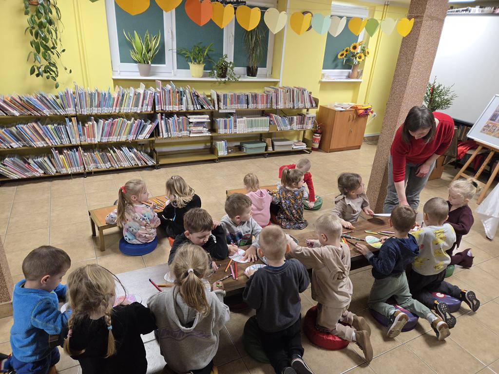 Szesnaścioro dzieci w kolorowych ubraniach siedzi przy ławeczkach i koloruje pisanki. Bibliotekarka w czerwonej bluzce i szarych spodniach nachyla się nad dziećmi. W tle są regały z książkami.