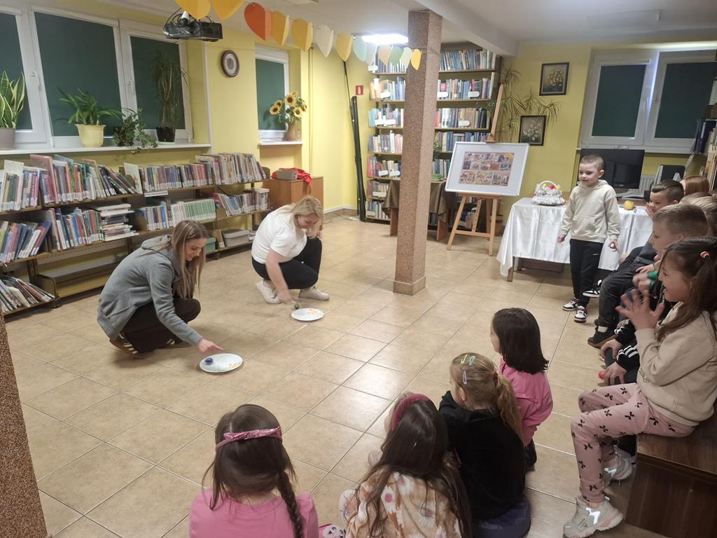 Dwie panie nauczycielki w kolorowych ubraniach kucają przed talerzami i kręcą pisanką. Po ich prawej stronie siedzi grupa dzieci w kolorowych ubraniach. W tle jest ekran od rzutnika.