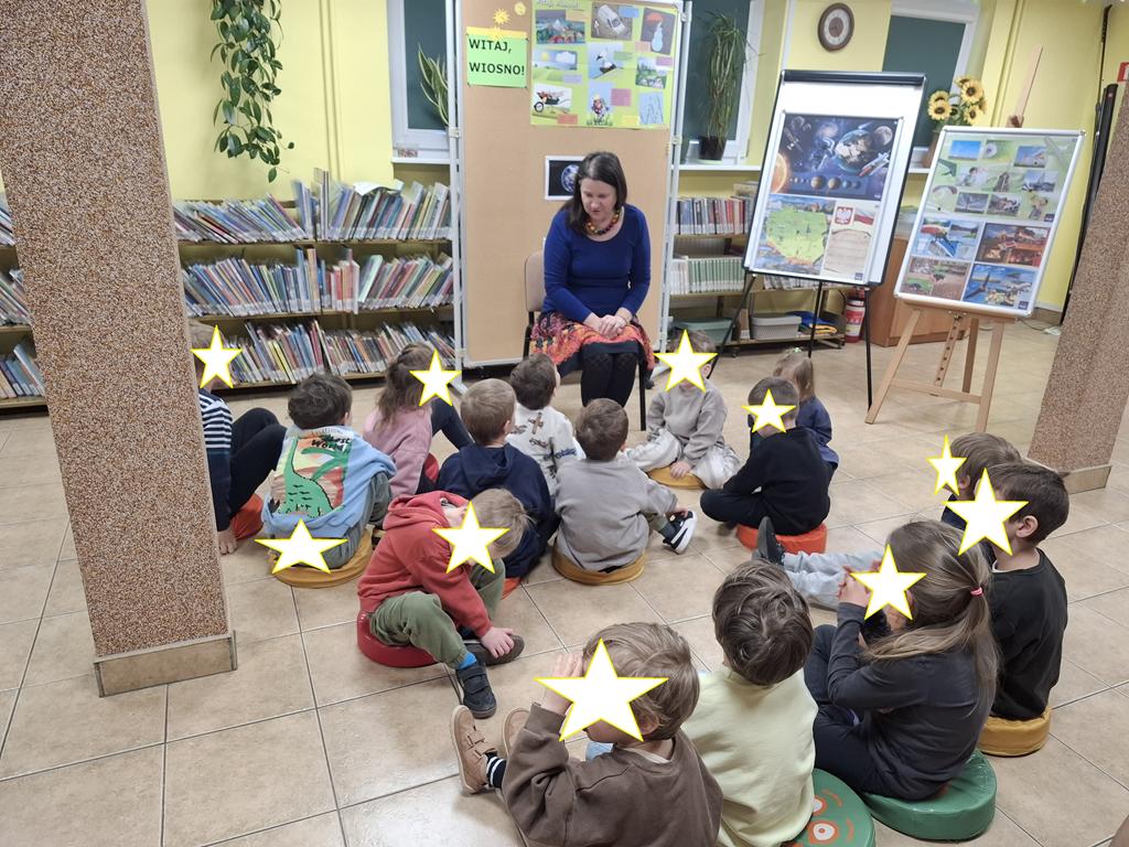 Kolorowo ubrana kobieta siedzi na krześle na środku sali. Za nią pod ścianą stoją niskie regały wypełnione książkami i tablice z kolorowymi ilustracjami. Przed nią na podłodze na siedziskach siedzi piętnaścioro dzieci w wieku przedszkolnym. Dzieci patrzą na kobietę.