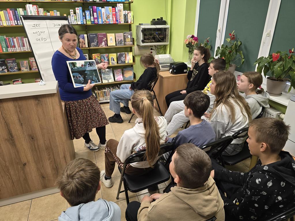 Kobieta stoi przy ladzie przodem do dzieci. W ręce trzyma kolorowe ilustracje. Dziesięcioro dzieci patrzy w stronę ilustracji. Za kobietą stoi tablica flipchart.