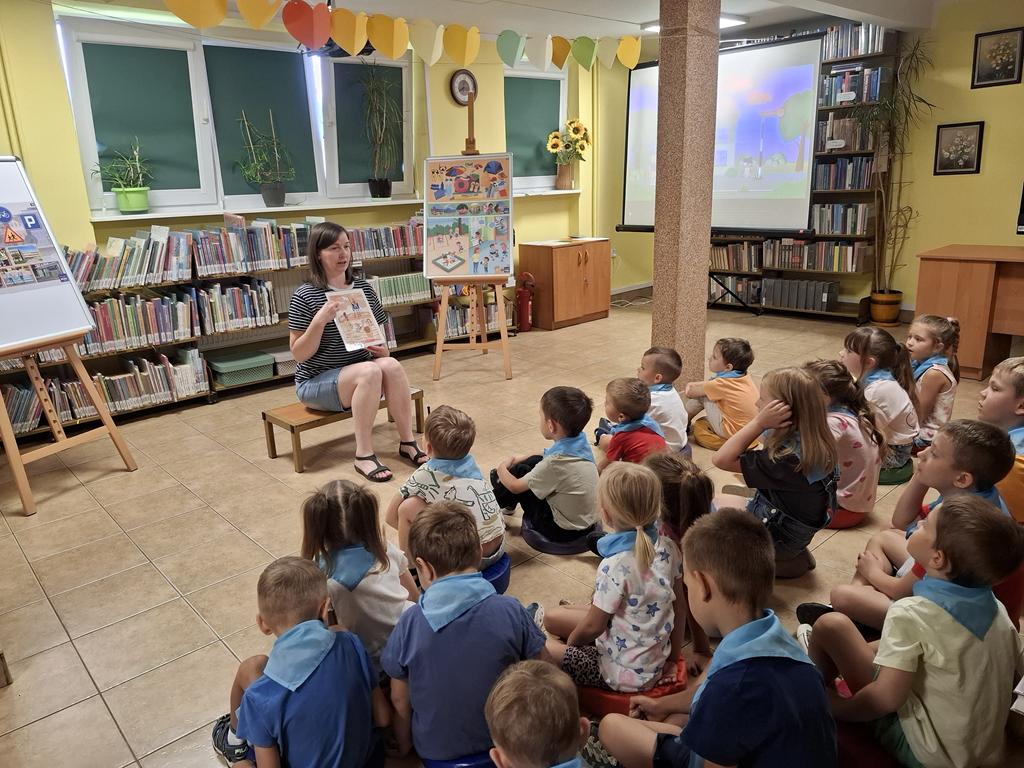 Bibliotekarka w bluzce w paski czarno-białe, niebieskich spodenkach siedzi na ławeczce i pokazuje dzieciom obrazki. Po jej prawej i lewej stroni stoją sztalugi z plakatami dotyczącymi bezpiecznych zachowań. Przed kobietą siedzi grupa przedszkolaków w kolorowych ubrania i niebieskich apaszkach. Po prawej stroni kobiety jest rzutnik ekranu. W tle są regały z książkami.