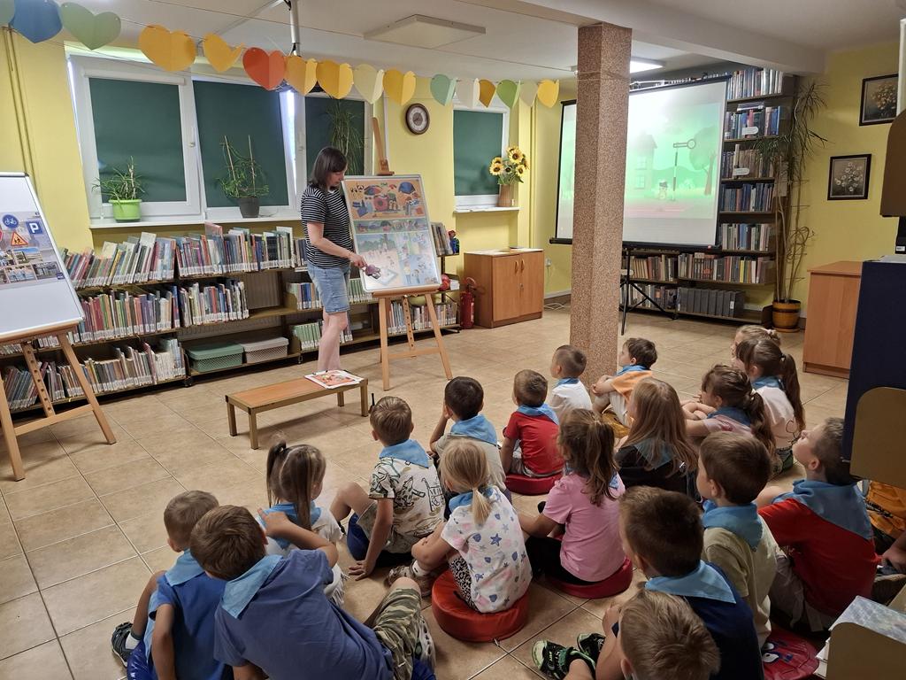 Bibliotekarka w bluzce w paski czarno-białe, niebieskich spodenkach stoi obok sztalugi z plakatami i wspólnie z dziećmi je omawia. Po jej lewej stroni stoi sztaluga z plakatami dotyczącymi bezpiecznych zachowań. Przed kobietą siedzi grupa przedszkolaków w kolorowych ubrania i niebieskich apaszkach. Po prawej stroni kobiety jest rzutnik ekranu. W tle są regały z książkami.