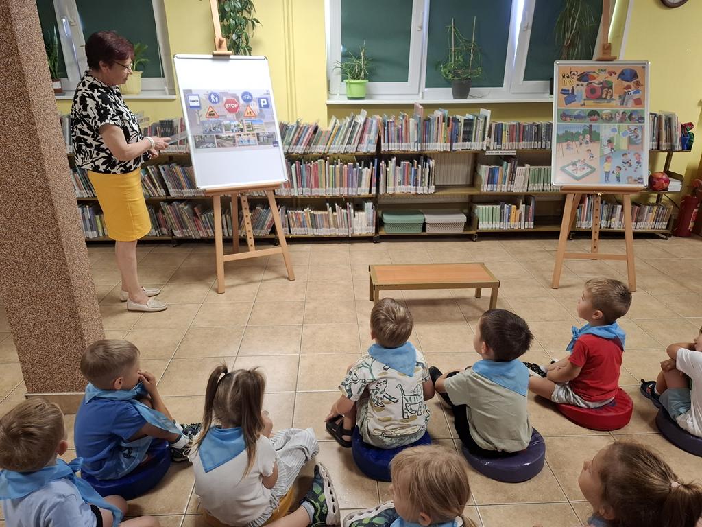 Bibliotekarka w w żółtej spódnicy i czarno-białej bluzce stoi przy sztaludze z plakatem i omawia go z dziećmi. Po jej prawej stroni jest też druga sztaluga z plakatami. Przed kobietą siedzi grupa przedszkolaków w kolorowych ubraniach i niebieskich apaszkach. W tle są regały z książkami.