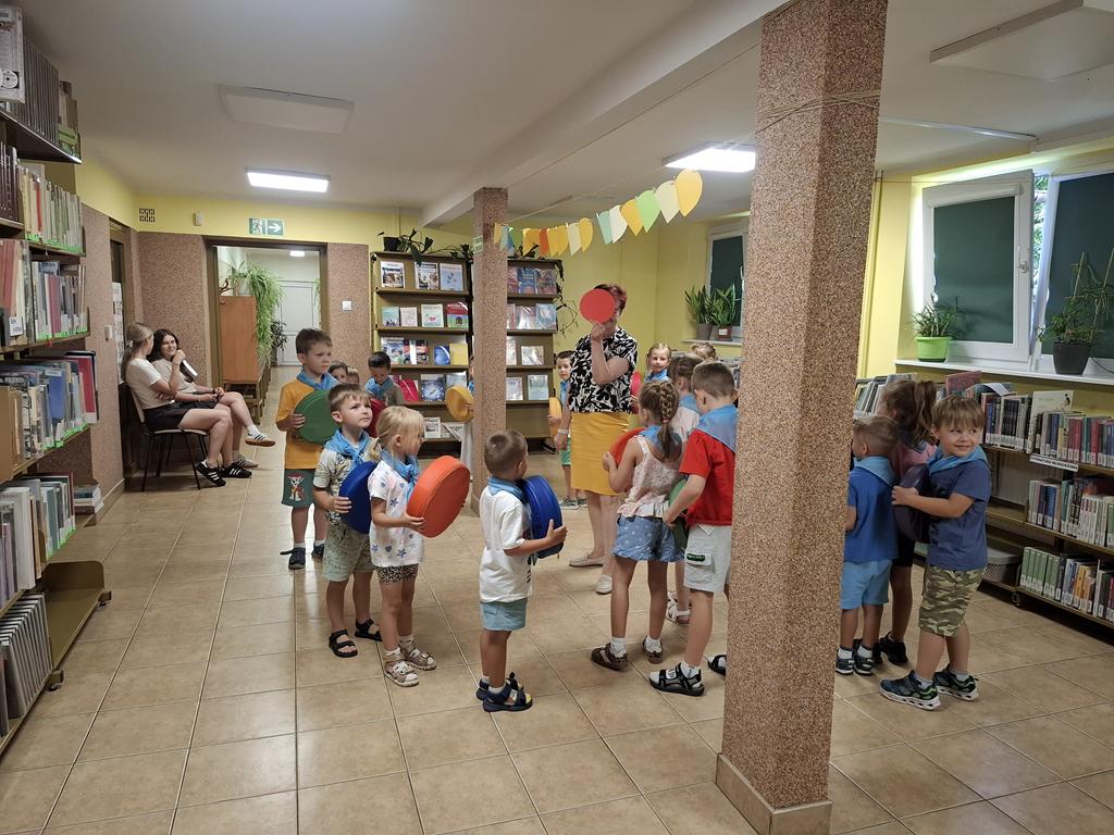 Grupa przedszkolaków w kolorowych ubraniach i niebieskich apaszkach stoi w okręgu. Dzieci trzymają kolorowe pufki. Na środku kręgu stoi bibliotekarka w żółtej spódnicy i czarno-białej bluzce trzymająca w ręku czerwone koło. W tle są regały z książkami.